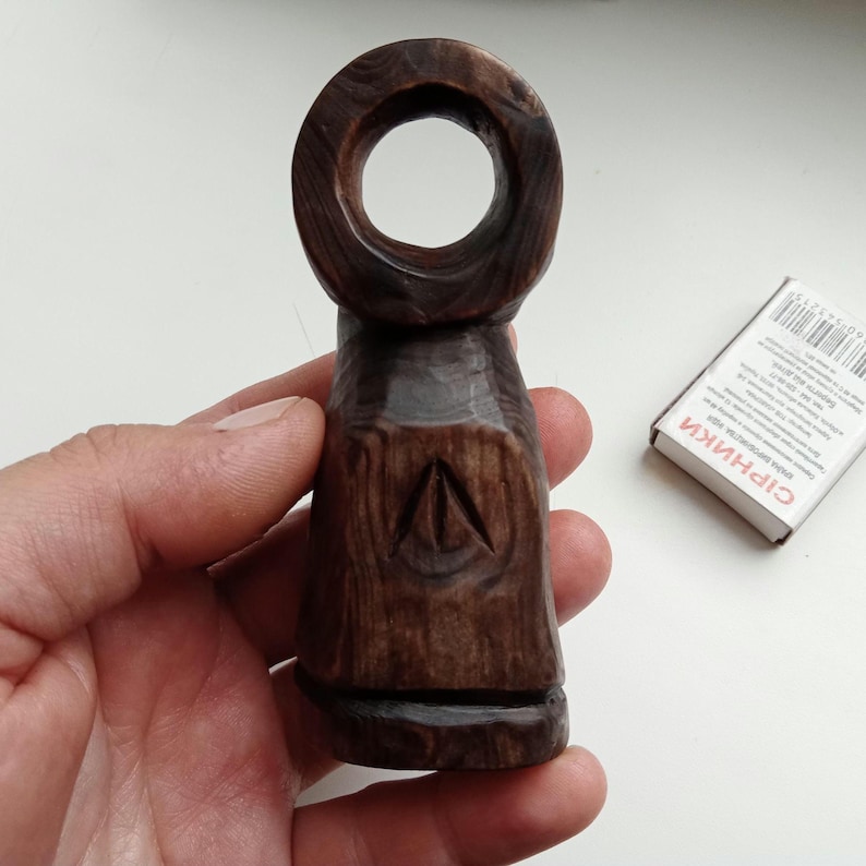 Hand holding a small, dark wooden figurine with a circular top and a carved triangular symbol on its body, likely a Slavic Panka doll or amulet.