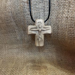 May include: A carved wooden cross pendant on a black cord. The cross features a detailed carving of a figure with outstretched arms. The pendant hangs against a textured, tan fabric background.