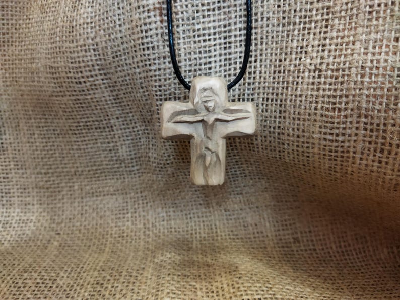 May include: A carved wooden cross pendant on a black cord. The cross features a detailed carving of a figure with outstretched arms. The pendant hangs against a textured, tan fabric background.