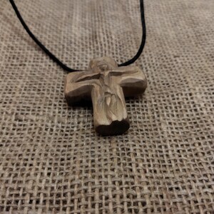 May include: A handcrafted wooden cross pendant on a black cord. The cross features a carved figure and is made of light brown wood. The necklace is displayed on a textured, tan fabric background.