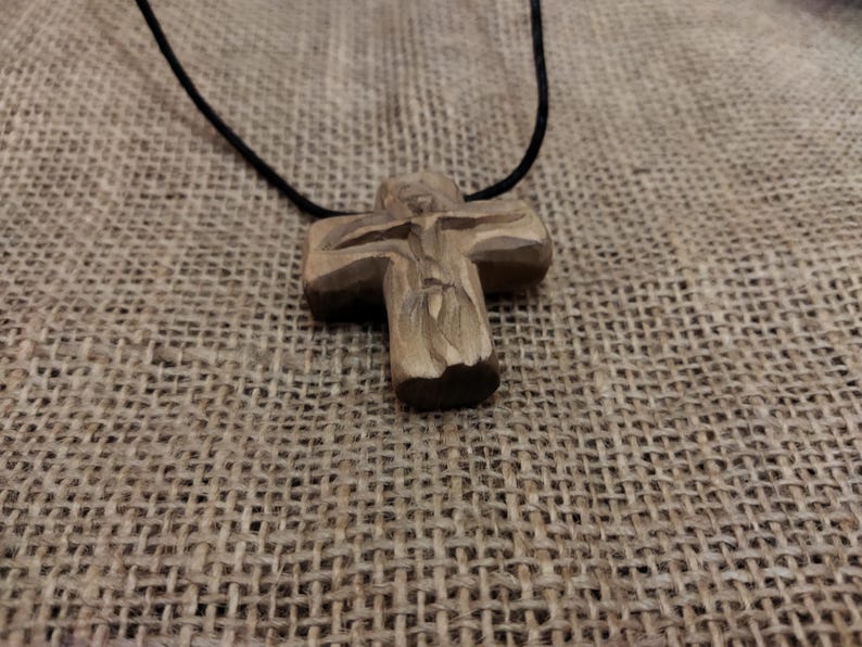 May include: A handcrafted wooden cross pendant on a black cord. The cross features a carved figure and is made of light brown wood. The necklace is displayed on a textured, tan fabric background.