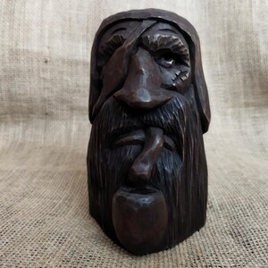 May include: A dark brown, carved wooden bust of a bearded man with an eye patch and a pipe. The sculpture has a weathered appearance, with detailed carving of the facial features and hair. The bust is set against a neutral background.