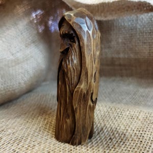 A 3.6-inch tall hand-carved linden wood figurine of a forest spirit. The figure has a long, flowing beard with deep textures and a face partially hidden under a hood. The dark brown stain highlights the artisan's hand-cut marks. This unique piece of Ukrainian folk art has a matte beeswax finish and a rustic, mystical appearance, shown against a neutral background.
