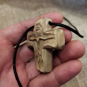 May include: A carved wooden cross pendant with a black cord. The cross features a detailed carving of a figure and is made of light-colored wood. The cord is approximately 18 inches long.