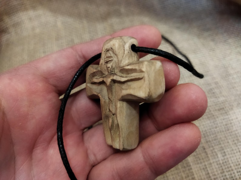 May include: A carved wooden cross pendant with a black cord. The cross features a detailed carving of a figure and is made of light-colored wood. The cord is approximately 18 inches long.