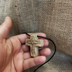 May include: A carved wooden cross pendant on a black cord. The cross features a detailed carving of a figure and is held in a hand. The wood has a natural, light brown color. The background is a textured, tan fabric.