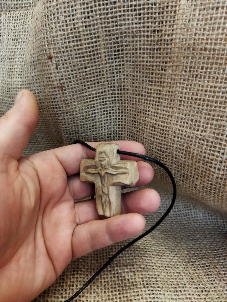 May include: A carved wooden cross pendant on a black cord. The cross features a detailed carving of a figure and is held in a hand. The wood has a natural, light brown color. The background is a textured, tan fabric.