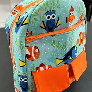 May include: A turquoise blue backpack with orange accents features a repeating pattern of cartoon fish, including Dory and Nemo. The backpack has two orange zippered pockets on the front.
