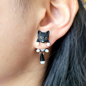May include: Black cat earrings with white accents. The cat's head is positioned on the ear, with the body and tail dangling below. The cat's eyes are yellow, and the paws are white. The earrings are a unique accessory.