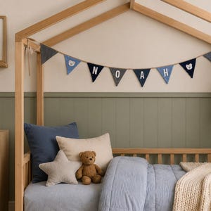 May include: A fabric bunting banner featuring triangular flags in shades of blue and grey, spelling out the name NOAH with white letters, interspersed with small bear face icons, hanging from a wooden house-shaped bed frame.