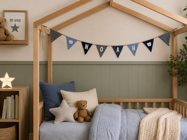 May include: A fabric bunting banner featuring triangular flags in shades of blue and grey, spelling out the name NOAH with white letters, interspersed with small bear face icons, hanging from a wooden house-shaped bed frame.