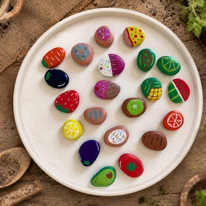 May include: A collection of twenty-one smooth, oval-shaped stones painted with colourful designs of various fruits, vegetables, and desserts, including strawberries, citrus slices, kiwi, aubergine, avocado, and sprinkled doughnuts, arranged on a white plate.