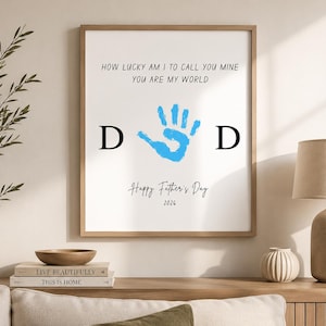 May include: A framed wall art print featuring the black text HOW LUCKY AM I TO CALL YOU MINE YOU ARE MY WORLD, a central blue handprint, the black letters D and D on either side, and the black text Happy Father's Day 2026 at the bottom.