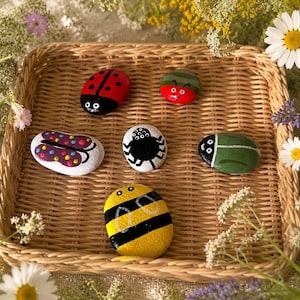 May include: Six smooth, oval-shaped stones painted with insect designs, including a red ladybug, a green beetle, a white butterfly with colourful dots, a black spider, a green bug, and a yellow and black striped bee, arranged in a woven basket.
