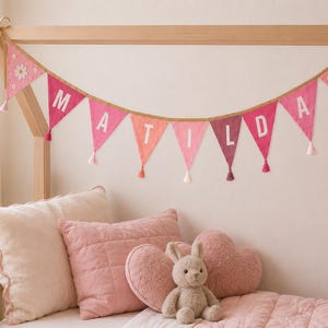 May include: A fabric bunting banner featuring triangular flags in shades of pink and red, spelling the name MATILDA in white letters. Each flag is accented with a small white tassel, and the end flags feature white daisy flower appliqu&eacute;s.
