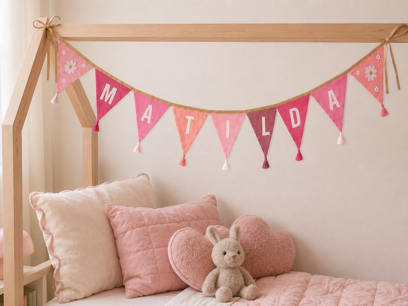 May include: A fabric bunting banner featuring triangular flags in shades of pink and red, spelling the name MATILDA in white letters. Each flag is accented with a small white tassel, and the end flags feature white daisy flower appliqu&eacute;s.