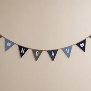 May include: A fabric bunting banner featuring triangular flags in shades of navy blue, grey, and light blue. The central flags display the white letters N, O, A, H, while two flags feature white bear face appliques on a blue background.