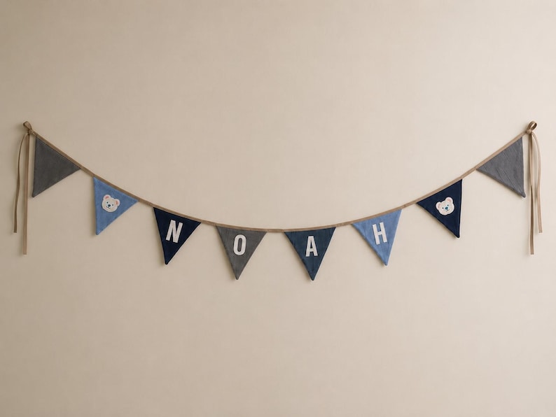 May include: A fabric bunting banner featuring triangular flags in shades of navy blue, grey, and light blue. The central flags display the white letters N, O, A, H, while two flags feature white bear face appliques on a blue background.