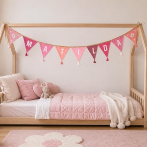 May include: A light wood house-shaped bed frame featuring a pink fabric bunting banner with white letters spelling MATILDA. The bed is dressed with a light pink quilted blanket, a white throw with pom-poms, and several pink pillows.
