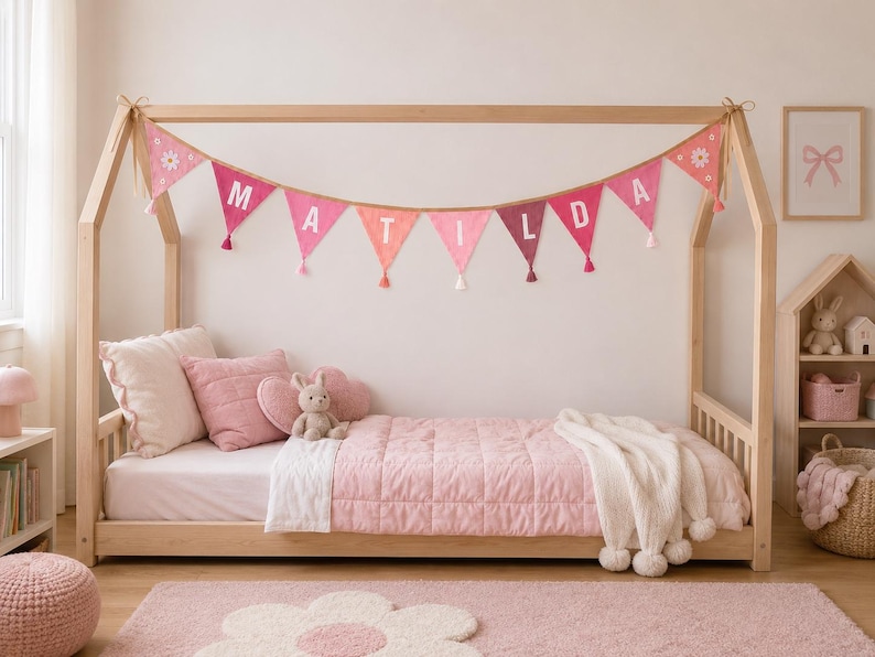 May include: A light wood house-shaped bed frame featuring a pink fabric bunting banner with white letters spelling MATILDA. The bed is dressed with a light pink quilted blanket, a white throw with pom-poms, and several pink pillows.