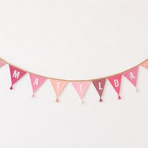 May include: A fabric bunting banner featuring triangular flags in various shades of pink, suspended on a tan string. The central flags display the white letters M, A, T, I, L, D, A, while the end flags are decorated with white daisy motifs.