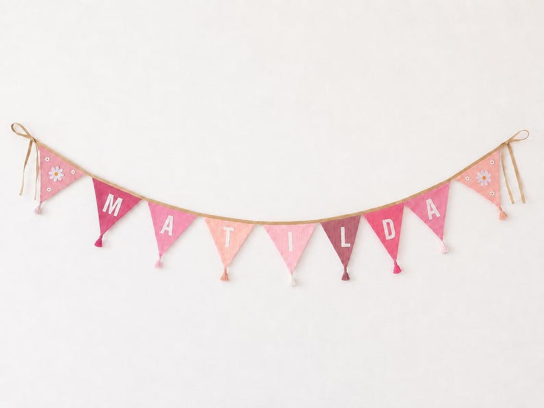 May include: A fabric bunting banner featuring triangular flags in various shades of pink, suspended on a tan string. The central flags display the white letters M, A, T, I, L, D, A, while the end flags are decorated with white daisy motifs.