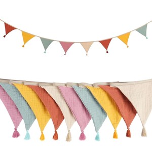 May include: A decorative bunting banner featuring triangular fabric flags in shades of pink, blue, yellow, and terracotta, each finished with a small matching tassel at the bottom point, suspended from a neutral-colored fabric cord.