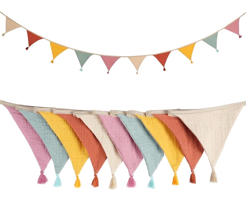 May include: A decorative bunting banner featuring triangular fabric flags in shades of pink, blue, yellow, and terracotta, each finished with a small matching tassel at the bottom point, suspended from a neutral-colored fabric cord.