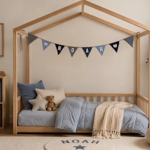 May include: A wooden house-shaped bed frame featuring a blue and grey fabric bunting with the name NOAH. The bed is dressed in a light blue quilted comforter, a cream throw blanket, a blue pillow, a star-shaped pillow, and a teddy bear.