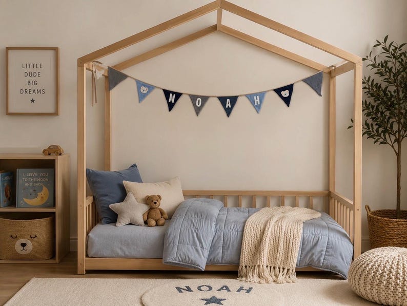 May include: A wooden house-shaped bed frame featuring a blue and grey fabric bunting with the name NOAH. The bed is dressed in a light blue quilted comforter, a cream throw blanket, a blue pillow, a star-shaped pillow, and a teddy bear.