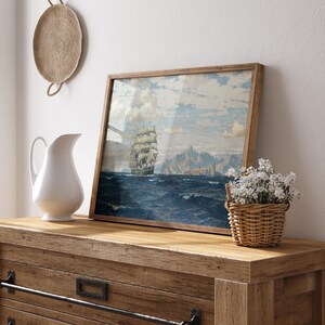 May include: A framed nautical painting of a tall ship sailing on a blue ocean under a cloudy sky. The artwork is in a wooden frame and rests on a wooden dresser. A white jug and a small basket of white flowers are also on the dresser.