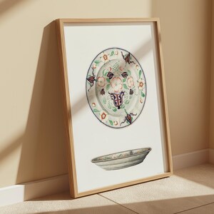 May include: A framed print of a vintage ceramic plate with a floral design in shades of blue, green, and white. The plate is surrounded by a white border and is displayed on a white background.