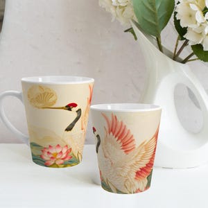 May include: Two white ceramic mugs with handles. Both mugs feature a colorful illustration of a crane with red, pink, and yellow accents. One mug features a crane with its wings spread, while the other mug features a crane with its wings folded. Both mugs have a white background with a light brown design.