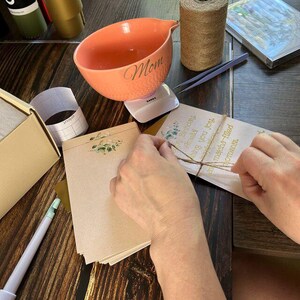 May include: An orange ceramic bowl with the word "Mom" in script, a spool of twine, and stationery with floral accents. A person is tying twine around a package with the words "Happy Birthday, matcha-filled moments."