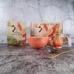 May include: A matcha tea set with a coral-colored bowl, bamboo whisk, sifter, and spoon. The set includes a recipe guide and a box, both featuring a crane illustration with red and white wings. The bowl has the word "Mom" on it.