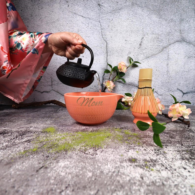 May include: A black cast iron teapot pours water into a peach-colored bowl with the word "Mom" inscribed on it. A matcha whisk and flowers are also present, suggesting a tea ceremony. Green matcha powder is scattered on the surface.