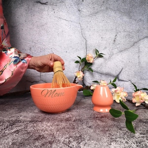 May include: A peach-colored matcha bowl with the word "Mom" etched on the side, a bamboo whisk, and a matching tea cup. The whisk is held above the bowl. Floral accents and a textured background complete the scene.