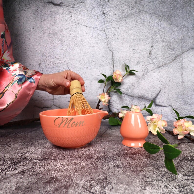 May include: A peach-colored matcha bowl with the word "Mom" etched on the side, a bamboo whisk, and a matching tea cup. The whisk is held above the bowl. Floral accents and a textured background complete the scene.