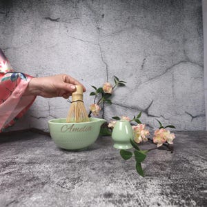May include: A light green matcha bowl with the name "Amelia" in gold lettering, a bamboo whisk, and a small matcha tea cup. The bowl has a textured pattern. Pink and white flowers and green leaves are in the background.