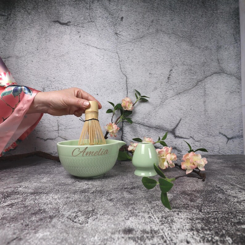 May include: A light green matcha bowl with the name "Amelia" in gold lettering, a bamboo whisk, and a small matcha tea cup. The bowl has a textured pattern. Pink and white flowers and green leaves are in the background.