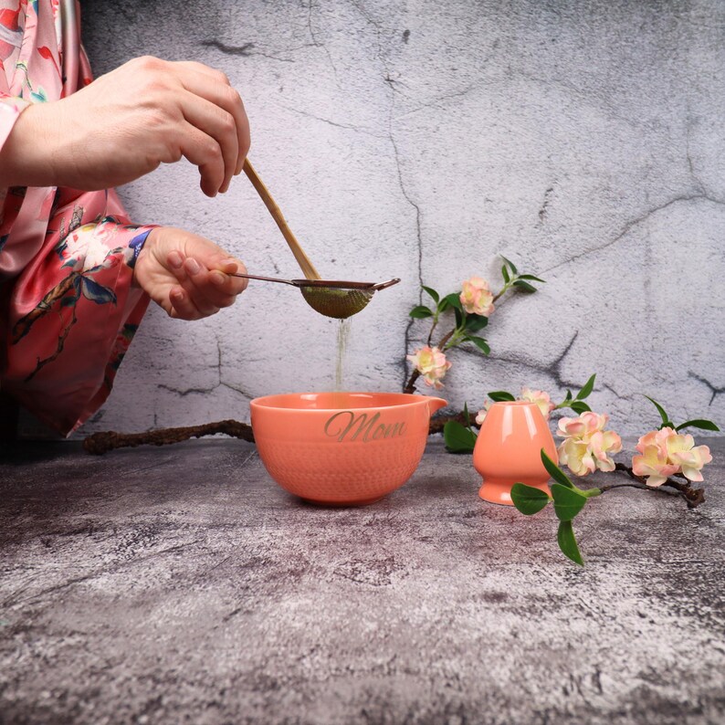 May include: A peach-colored ceramic bowl with the word "Mom" in cursive, a small matching pitcher, and a tea strainer. A wooden whisk is held over the bowl, with liquid pouring through the strainer. Decorative flowers and branches are in the background.