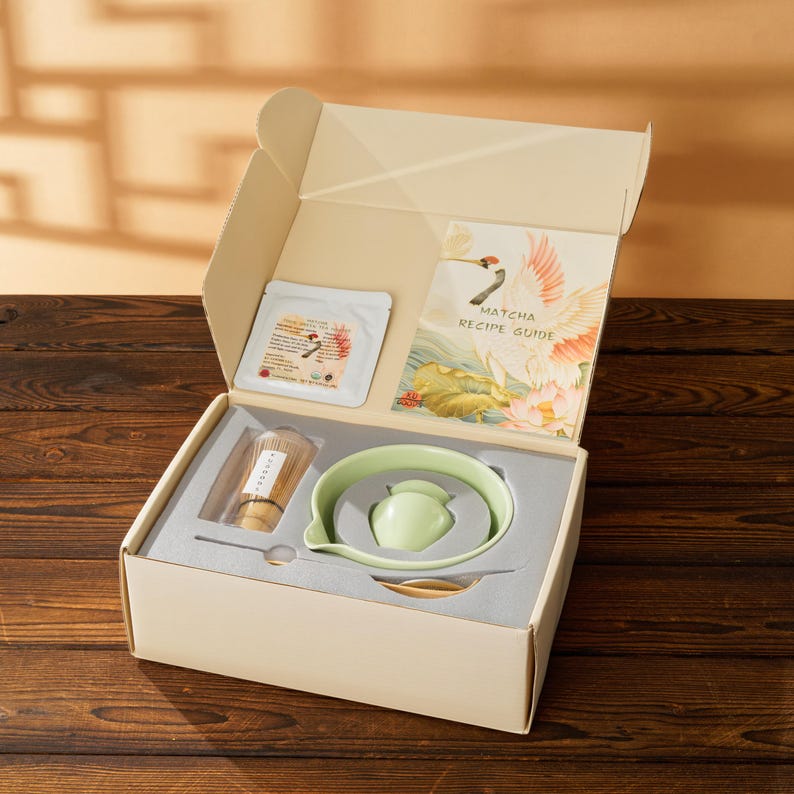 May include: An open gift box containing matcha tea set components. The set includes a green ceramic bowl, whisk, spoon, and a matcha recipe guide. The box also contains a packet of matcha tea and a bamboo whisk.