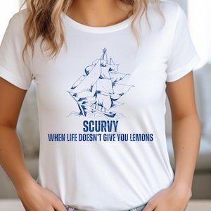 May include: White t-shirt with a blue illustration of a sailing ship and the text "SCURVY WHEN LIFE DOESN'T GIVE YOU LEMONS". The shirt is made of a soft material and has short sleeves.