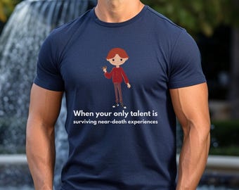 May include: A navy blue t-shirt with a cartoon illustration of a person with red hair holding a small object. The text on the shirt reads "When your only talent is surviving near-death experiences."