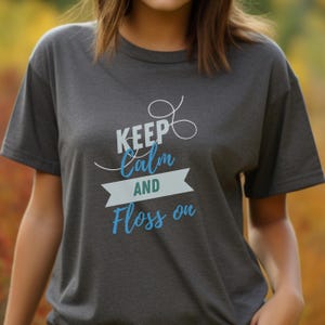 Könnte beinhalten: Ein dunkelgraues T-Shirt mit dem Text "Keep Calm and Floss On" in weißer und blauer Schrift.