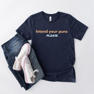 May include: A navy blue t-shirt with the text "Intend your puns PLEASE" in orange and light blue lettering. The shirt is paired with blue jeans and white sneakers.