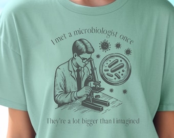 May include: Sage green t-shirt featuring a black and white illustration of a person using a microscope. The text reads, "I met a microbiologist once. They're a lot bigger than I imagined."