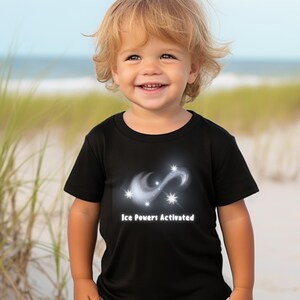 May include: A black t-shirt with a white graphic of a swirling galaxy with stars and the text "Ice Powers Activated". A young child is wearing the shirt and smiling.