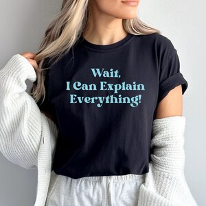 Op de afbeelding: Zwart T-shirt met de tekst "Wait, I Can Explain Everything!" in lichtblauw. Het shirt heeft korte mouwen en een ronde hals. De tekst staat in het midden van de voorkant van het shirt.