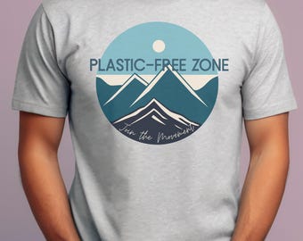 May include: A light gray t-shirt featuring a circular graphic with a mountain range design in shades of blue and white. The text "PLASTIC-FREE ZONE" is above the mountains, and "Join the Movement" is below.